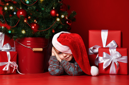 Happy xmas and New Year. Portrait of child in Santa red hat waiting for Christmas gifts. Little baby boy lies under the Christmas tree with presents and is waiting for a miracleの写真素材
