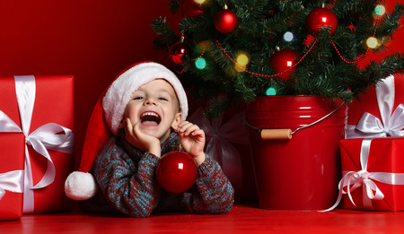 Happy xmas and New Year. Portrait of child in Santa red hat waiting for Christmas gifts. Little toddler boy lies under the Christmas tree with gifts and holds the Christmas ball.の写真素材