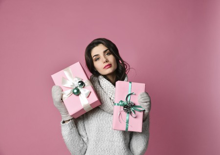 Cheerful excited woman in sweater holding two gift boxes and looking at camera isolated on pink...
