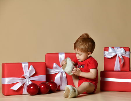Portrait of a baby dressed a red bodysuit with big red balls decorations on the xmas present gifts over beige nude studio background. Baby first christmas. Beautiful little baby celebrates Christmas.の写真素材