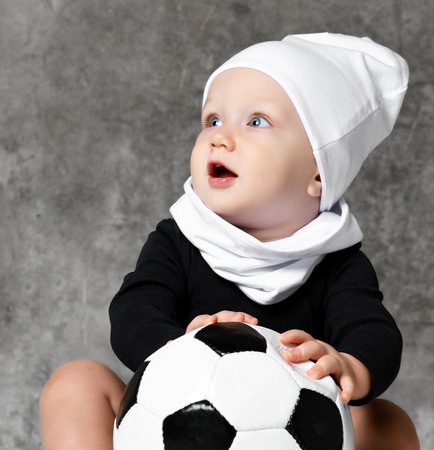little boy looks surprised aside, holds a black and white football in his hands. wearing a white cotton hat and a black bodysuitの写真素材