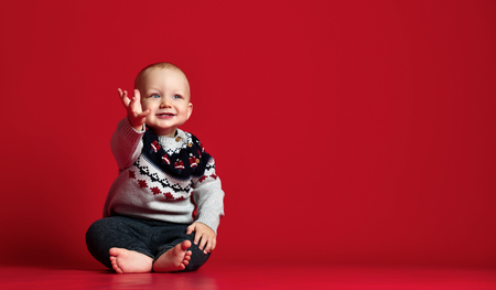 Image of sweet baby boy, closeup portrait of child, cute toddler in sweater with blue eyes.small boy sits, smiles and pulls hand up over red studio backgroundの写真素材