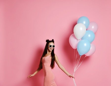 Smiling beautiful young woman posing with pink and blue balloons on pink background, studio shot. Have a fun birthday party happy and sexyの写真素材