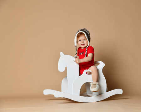 little cute red-haired toddler boy at warm hat sits astride a white rocking horse  studio shot, happy baby. smilesの写真素材
