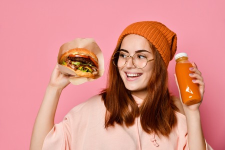 Beautiful teenage girl with red hair and hat holding burger and beverage in both hands. Ginger student girl has fast food lunchの写真素材