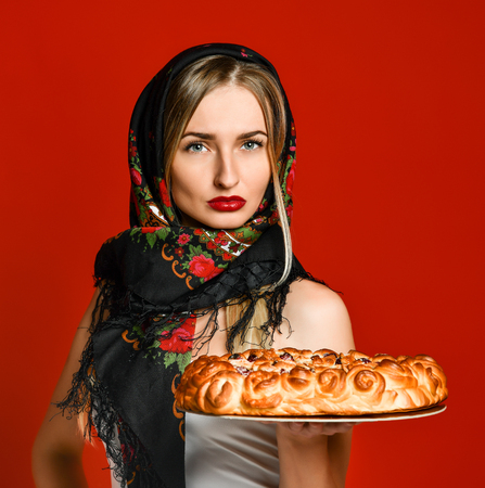 Portrait of a young beautiful blonde in headscarf holding a delicious homemade cherry pie over red backgroundの写真素材