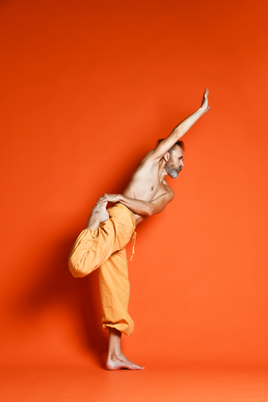 Close up of old man practicing yoga, doing Natarajasana exercise, Lord of the Dance pose, working out, wearing orange pants, top, indoor full length, studioの写真素材