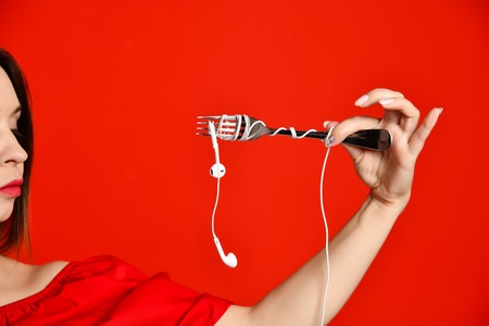 Attractive girl in red dress holding a fork in hands. on the headphone plug. prepared to eat. Music saturationの写真素材