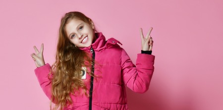 portrait of a little fashionable girl in a warm pink down jacket and jeans, looks into the camera and shows Victoria a cool gesture with her fingers, on a pink backgroundの写真素材