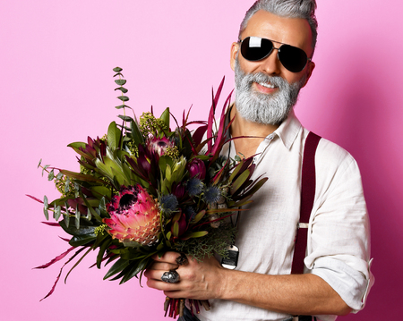 Handsome older stylish bearded man in white shirt holding a bouquet of flowers over pink background and looking at camera in sunglassesの写真素材
