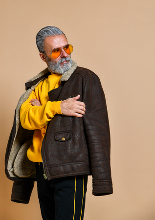 Portrait of senior stylish rich man with a beard and mustache in a leather coat, a stern look away through yellow modern glasses, the studio on a beige backgroundの写真素材