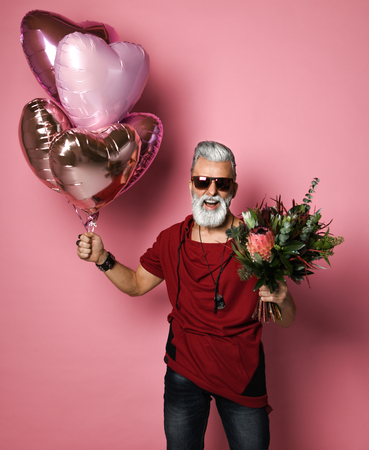 Stylish bearded middle-aged man with a modern hairstyle and fashionably dressed holding bunch of heart shaped balloons and a bouquet of flowers. Concept Valentine's Day, Dateの写真素材