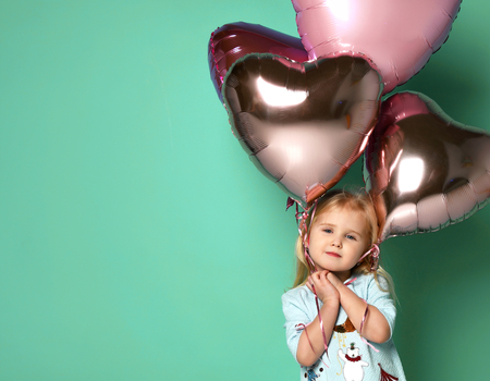 little girl has fun with a stack of pink air balloons in the shape of her heart. child holds balloons over the finished and looks through the ribbons into the cameraの写真素材