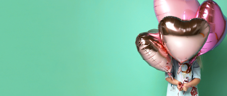 little girl has fun with a stack of pink air balloons in the shape of her heart. child hid behind a group of balloonsの写真素材