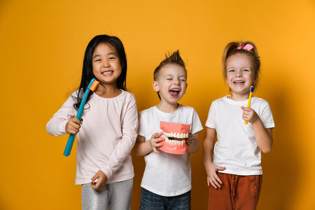 Three children with toothbrushes and a kissuos mock-up with zooms stand over yellow background wall and smile rejoicing at the camera.The concept of health, oral hygiene, people and beauty.の写真素材