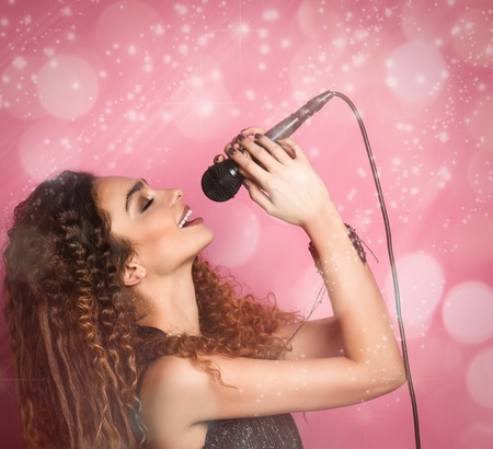 Young beautiful curly singer beauty sings into the microphone alone, against the background of pink bokehの写真素材