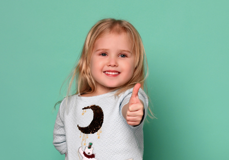Little adorable blonde girl in a blue dress on the background shows a thumbs up in agreement! Indoor shotの写真素材