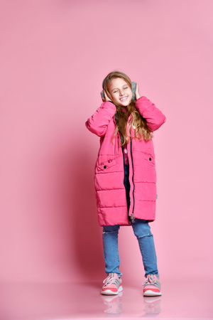 full-length portrait of Cute smiling girl in winter puffer jacket and fur headphones over pink wall backgroundの写真素材