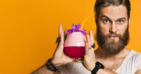 Attractive, stunning and cheerful bearded young man holding a multicolored cocktail with a straw and looking at the camera. On a yellow background.の写真素材