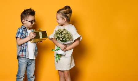 Picture of happy young couple boy and girl standing over yellow wall and looking aside. Boy gives a flowers and gift to girl. Concept friendship, loveの写真素材