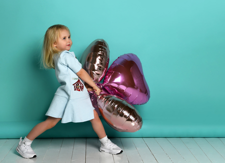 little girl has fun with a stack of pink air balloons in the shape of her heart. child holds the balls on the side and looks away.の写真素材