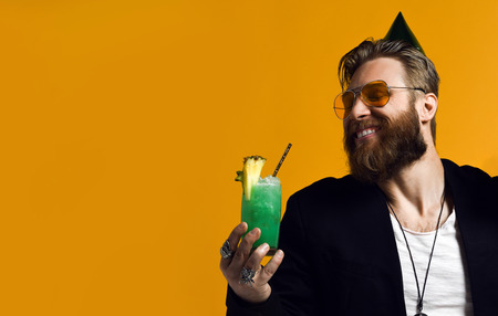 handsome bearded man with cone hat beard holding glass of cocktail and gift box celebrating birthday among friends isolated on yellow. Partyの写真素材