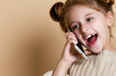 Little girl in oversized sweater happily chatting on by cell phone with his classmates nude beige backgroundの写真素材