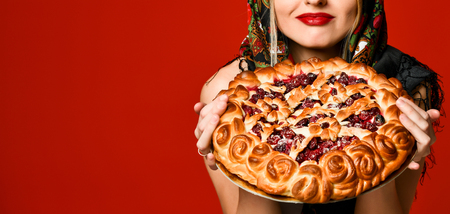 Portrait of a young beautiful blonde in headscarf holding a delicious round homemade berry pie over red backgroundの写真素材