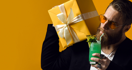 handsome bearded man with stylish hair beard tries to drink a cocktail and hold gift box isolated on yellow. Partyの写真素材