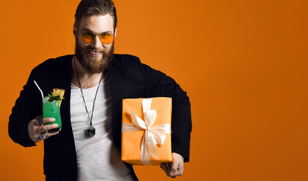 handsome bearded man with stylish hair beard holding glass of cocktail and gift box isolated on yellow. Partyの写真素材