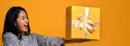 Portrait of a smiling cute asian girl holding a gift box , isolated over a yellow background. emotions of joy from receiving a giftの写真素材