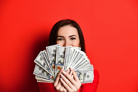 Young luxurious pretty woman in red dress hiding behind a pile of money banknotes fanned out looking...