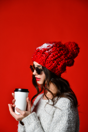 Happy pretty stylish young woman in sunglasses and knitted hat drinking coffee-to-go and thinking over red wallの写真素材