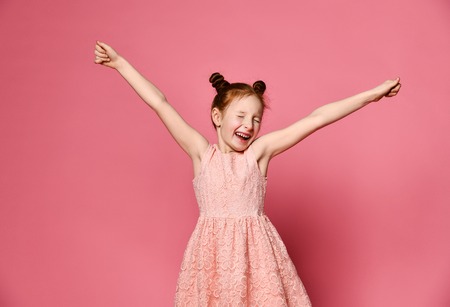 Portrait of a young girl with red hair in a cute dress smiling at the camera with her arms in the air, isolated on a bright pink backgroundの写真素材