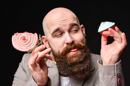 happy bearded bald man in a jacket holds two cream cakes on black background. A man squints between two sweetsの写真素材