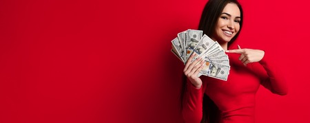 Young saucy brunette woman in red dress shows with a finger over a pile of dollars banknotes money in her hand, a place for advertising, isolated on a red backgroundの写真素材