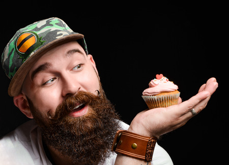 happy bearded man in a trendy stylish cap is holding two cream muffin cakes on a cobbler, making a choice of which one to eat. on a black background.の写真素材
