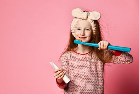 Portrait of a cute red-haired baby girl in pajamas and a hair band holds a toothbrush and toothpaste isolated on pink background. The concept of everyday oral hygiene.の写真素材