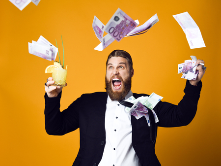 I am rich! Happy young businessman with glass of cocktail in formal clothes holding bunch of money...