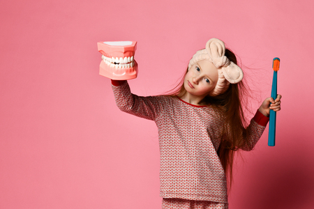 Red-haired pretty girl in pajamas with a toothbrush and a kissuos mock-up with scales stand over a pink background wall and the joy of a smile on camera.の写真素材