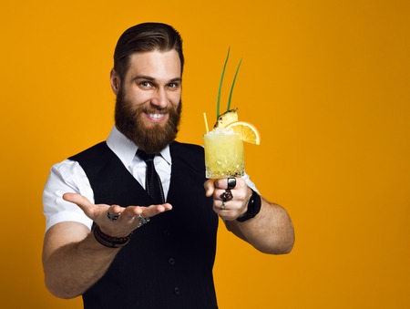 handsome bearded barman with a long beard and mustache has stylish hair offers to treat himself and try a cocktail of his cooking, he is wearing a classic waistcoat on a yellow studio backgroundの写真素材