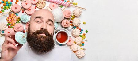 Happy bald bearded brutal man lies among marshmallow and candy sweets, holds a cake in his hand and is going to eat it with a cup of tea. Copy spaceの写真素材
