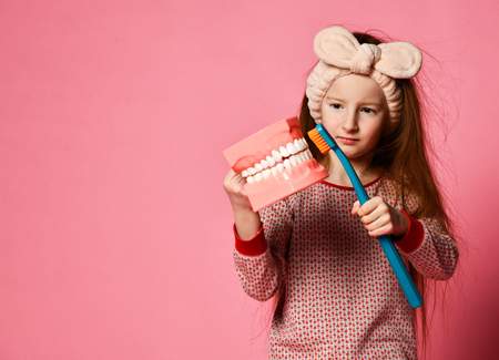 Red-haired pretty girl in pajamas with a toothbrush and a kissuos mock-up with scales stand over a pink background wall and the joy of a smile on camera.の写真素材