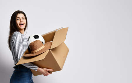 Young attractive brunette girl holding a cardboard box with things - plant, hat, ball and others. A woman is infinitely happy to move to her new dream home.の写真素材