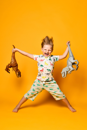 little cheerful boy holds a plastic dinosaur in each hand. The boy is dressed in a suit with pictures of dinosaurs, on yellow backgroundの写真素材
