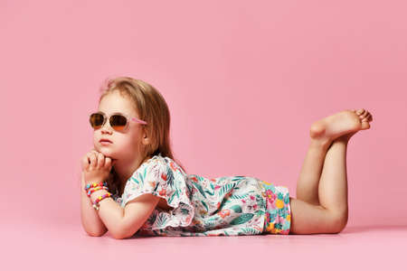Little girl child looks to the side lying on the floor of the studio in summer clothes in sunglasses on a pink background one.の写真素材