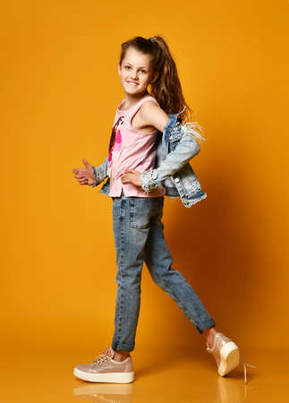 young teen girl model posing on a yellow background in jeans and a jacket, with long hair gathered in a high tail, stands half a turn to the camera taking off a jacket. Fashionable childhoodの写真素材