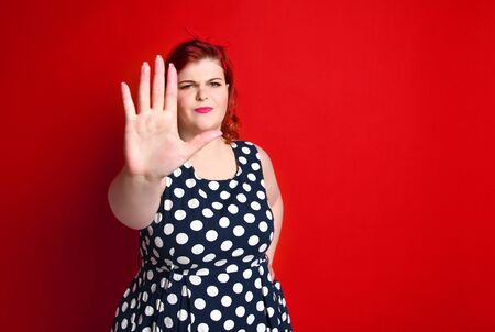 Serious young caucasian size woman plus showing stop gesture with her hand. She dressed in a polka dot dress in the studio on a red backgroundの写真素材