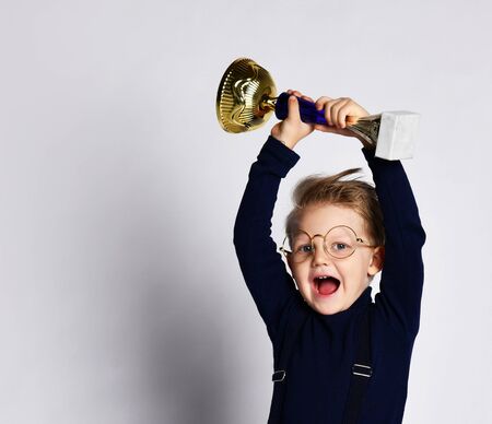 Excited smart little boy in jeans, sweater and glasses celebrates his golden trophy,の写真素材