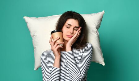 Tired happy woman drinking coffee at home or office having too much work. Bored businesswoman with a pillow and coffee cups. Sleep all day, late for workの写真素材